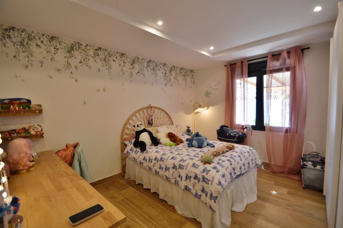 Chambre lumineuse avec tête de lit en rotin, mur décoré d'un motif floral et parquet. L'éclairage encastré au plafond complète cette atmosphère douce et moderne.