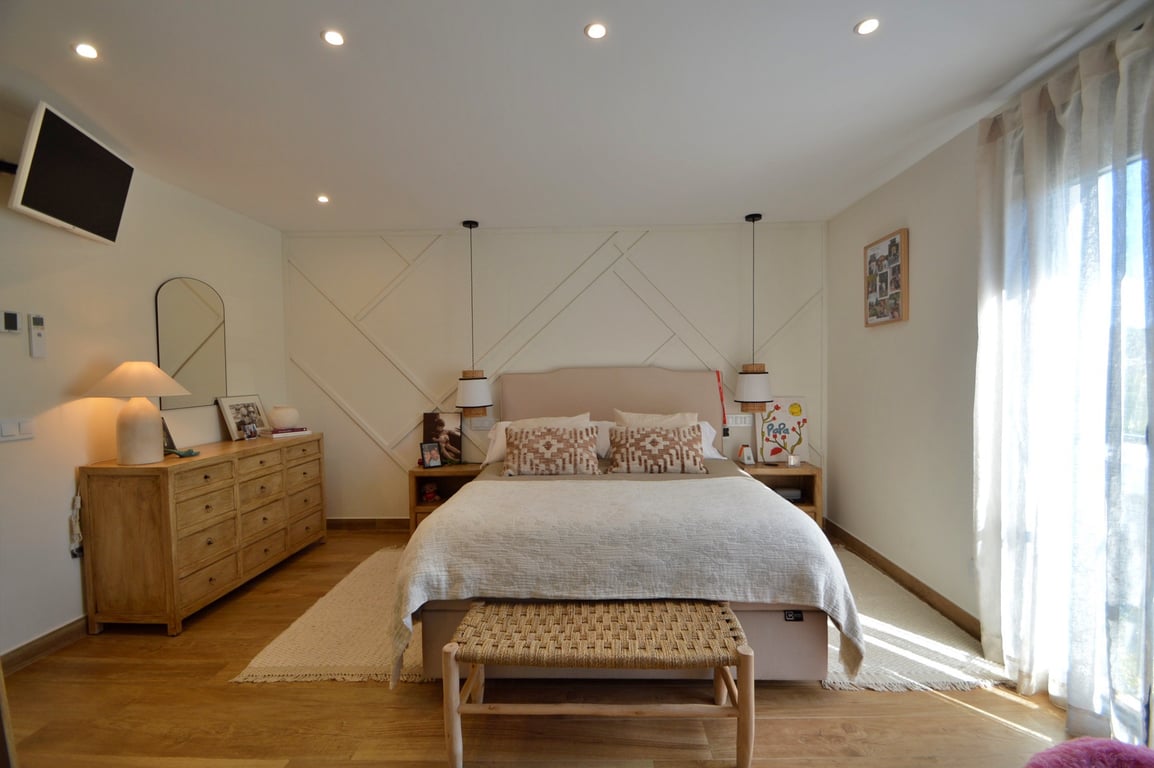 Chambre principale avec mur décoratif géométrique, parquet clair et éclairage encastré. Elle dispose d'une commode en bois, de luminaires suspendus et de rideaux vaporeux.