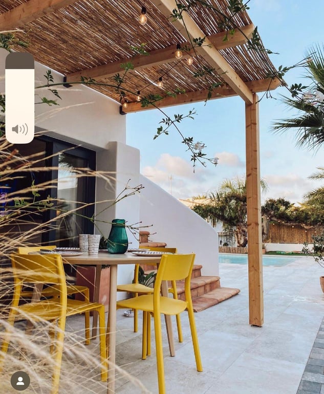 Coin repas extérieur sous une pergola en bois et canisse avec guirlandes. Chaises jaunes modernes sur une terrasse en pierre face au jardin et à la piscine.