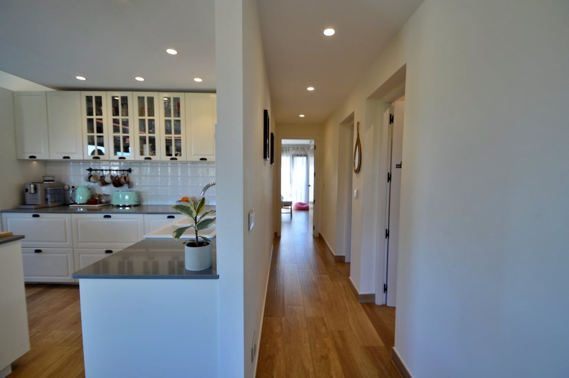 Couloir lumineux avec sol aspect bois menant à une cuisine moderne aux meubles blancs, plans de travail gris et éclairage encastré au plafond.