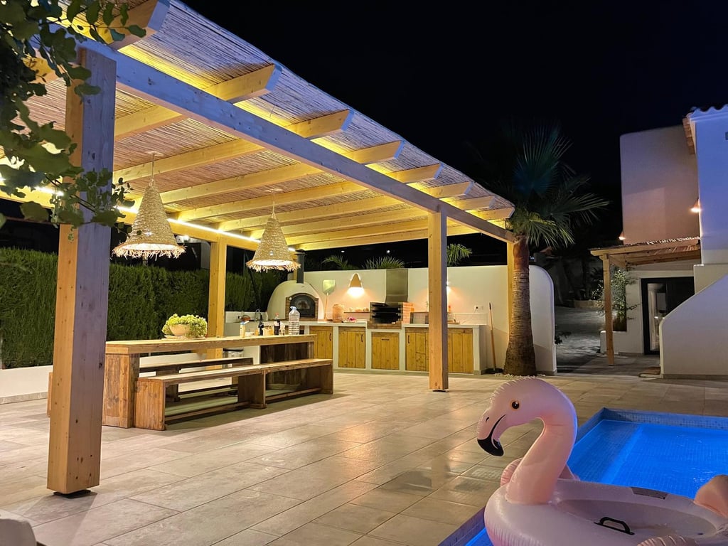 Cuisine d'été éclairée avec four à pizza, barbecue et espace dînatoire sous une pergola en bois, située en bordure de piscine.