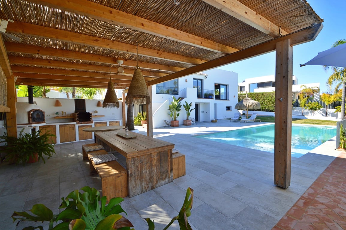 Cuisine d'été sous pergola en bois avec four à pizza, espace repas rustique et vue sur la piscine et la villa blanche de style Ibiza.