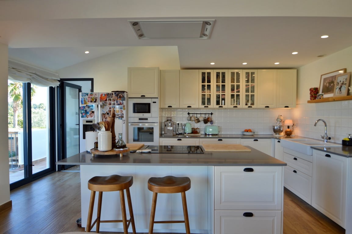 Cuisine moderne à aire ouverte avec îlot central, tabourets en bois et plans de travail gris. Elle dispose d'appareils intégrés et d'un accès direct vers l'extérieur.