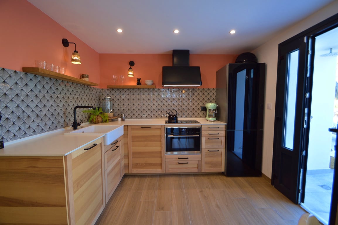 Cuisine moderne avec meubles en bois clair, évier à poser blanc et crédence géométrique. Les appareils noirs contrastent avec le mur terracotta et les étagères en bois massif.
