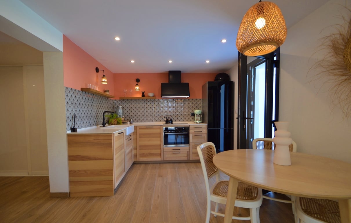 Cuisine moderne avec meubles en bois, mur d'accent terre cuite et coin repas comprenant une table ronde, des chaises en cannage et une suspension en fibres naturelles.