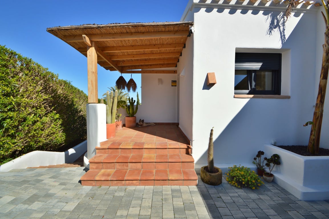Entrée de style ibizien avec pergola en bois et canisse, marches en terre cuite et cactus décoratifs devant une façade blanche épurée.