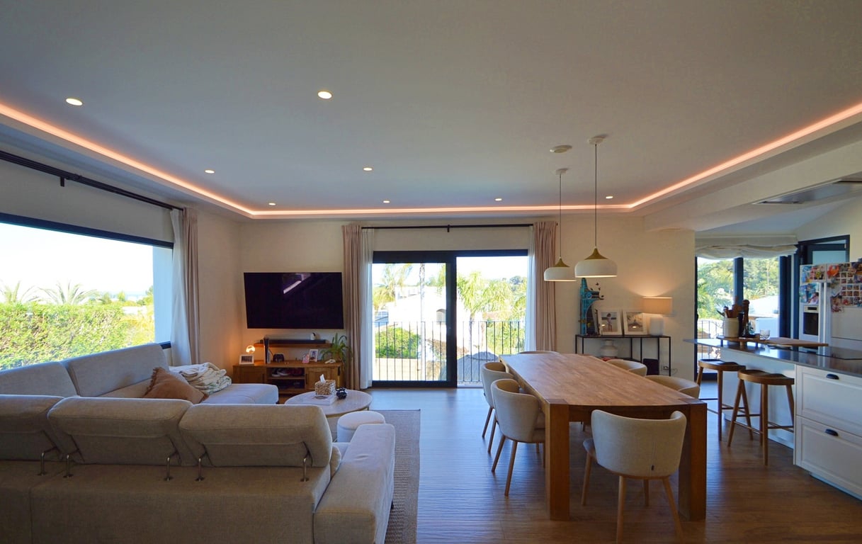 Espace de vie ouvert comprenant un salon lumineux et une salle à manger avec éclairage LED indirect, mobilier en bois et accès direct vers l'extérieur.