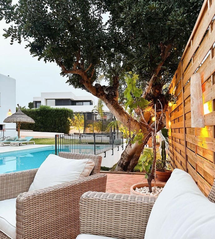 Espace détente extérieur avec fauteuils en rotin, sol en terre cuite et clôture en bois à éclairage chaleureux bordant la piscine et un arbre mature.