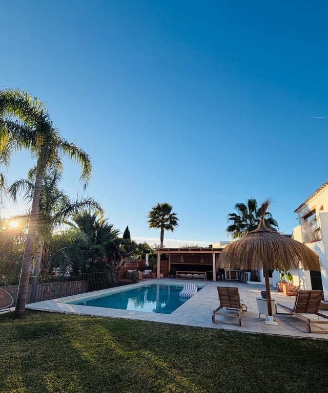 Espace piscine avec terrasse en pierre, parasol en brande et chaises longues. Cuisine d'été couverte et jardin méditerranéen avec palmiers sous un ciel dégagé de fin de journée.