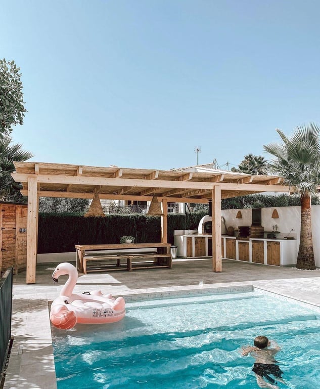 Espace repas extérieur et cuisine d'été avec four à pizza et barbecue, abrités par une pergola en bois face à la piscine au sel.