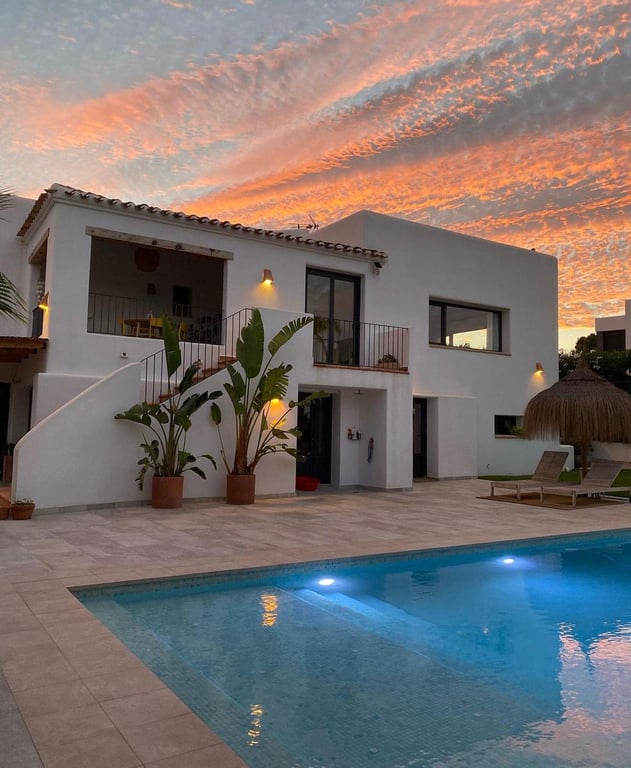 Extérieur de style ibizane au crépuscule avec piscine au sel éclairée, vaste terrasse en pierre et escalier menant à la naya sous un ciel flamboyant.