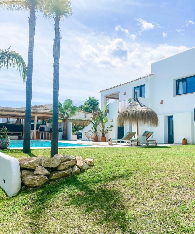 Extérieur de style ibizane avec pelouse, piscine et parasol en brande. En arrière-plan se trouve la cuisine d'été avec son four à pizza traditionnel.