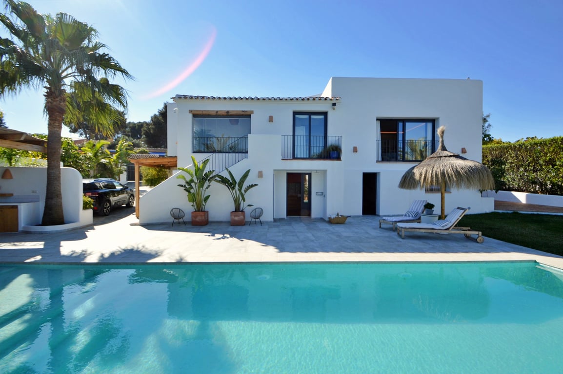 Façade blanche de style Ibiza face à une grande piscine au sel, avec terrasse en pierre, palmiers et espace de détente avec parasol en bruyère.
