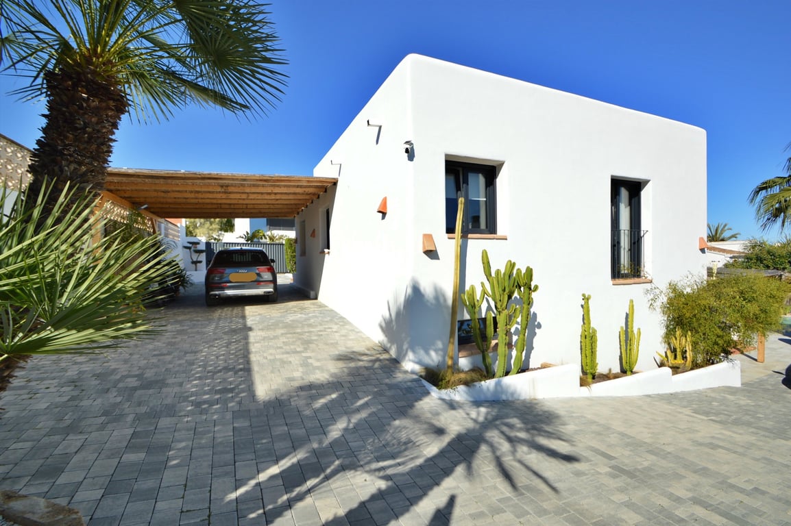 Façade blanche de style ibizane avec abri de voiture en bois, allée pavée et jardin méditerranéen agrémenté de cactus et palmiers.
