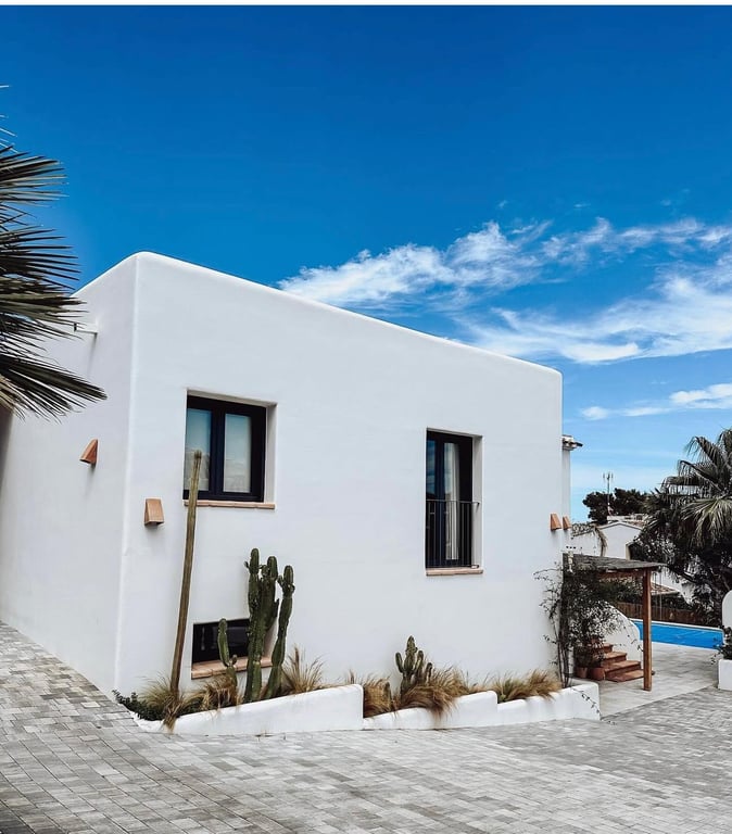 Façade de style ibizane aux murs blancs lisses, fenêtres à menuiserie sombre et jardin de cactus intégré bordant une allée pavée de pierres grises.