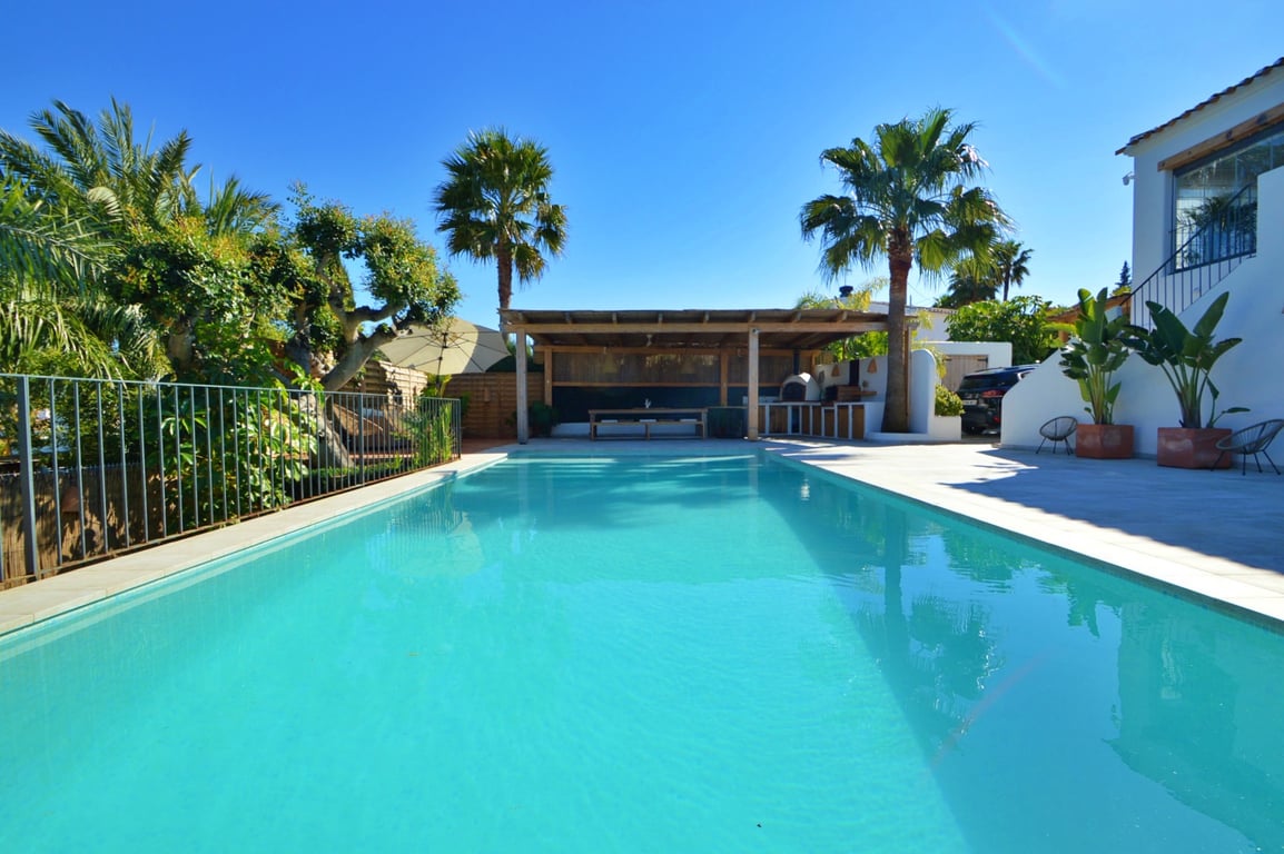 Grande piscine rectangulaire avec terrasse ensoleillée, comprenant un espace dînatoire sous pergola en bois et un jardin méditerranéen arboré de palmiers.
