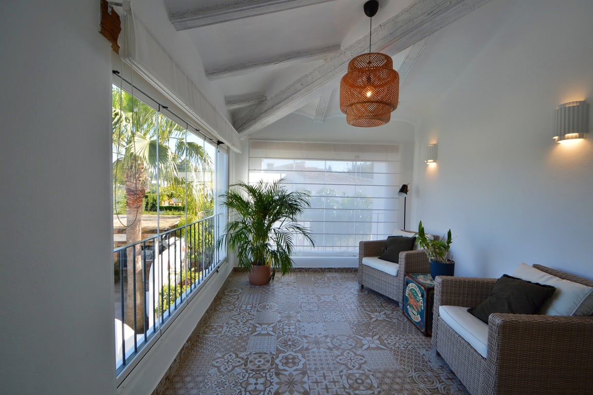 Naya vitrée avec poutres en bois blanchi, sol en carreaux de ciment décoratifs et fauteuils en osier donnant sur le jardin extérieur.
