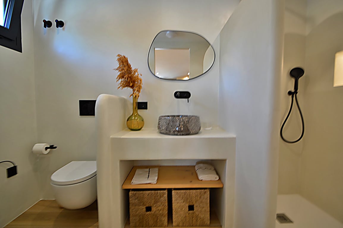 Salle de bain de style Ibiza avec murs en microciment, vasque en pierre et robinetterie noire mate. Elle dispose d'un miroir organique et d'un meuble vasque maçonné avec rangements.