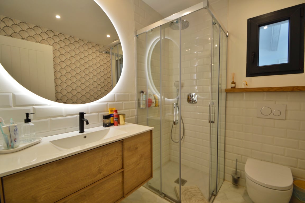 Salle de bain moderne avec miroir circulaire rétroéclairé, meuble en bois et douche d'angle revêtue de carreaux de métro blancs.