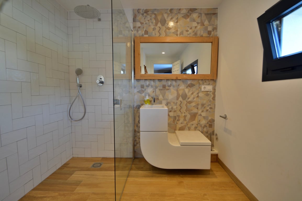 Salle de bains moderne avec douche à l'italienne, paroi vitrée et combiné vasque-WC design. Elle dispose d'un mur en carreaux géométriques et d'un miroir à cadre en bois.