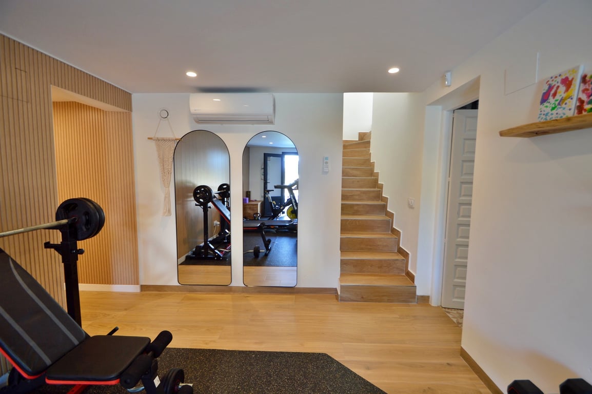Salle de sport privée avec miroirs en arche, sol imitation bois, climatisation et escalier intégré, offrant un espace d'entraînement moderne et lumineux.