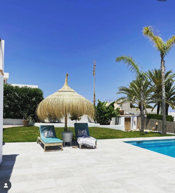 Terrasse en pierre claire avec transats turquoise et parasol en paille bordant la piscine, au cœur d'un jardin avec pelouse et palmiers.