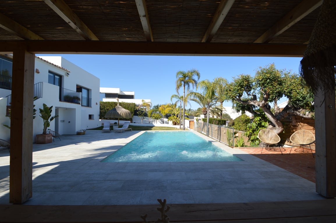 Vue depuis la pergola en bois sur la piscine au sel, entourée de terrasses en pierre claire, de palmiers et de la façade blanche de style ibizien.