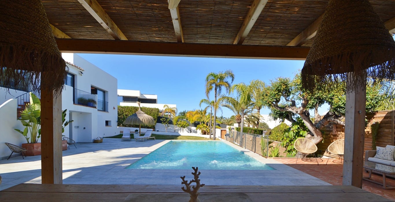Vue depuis la terrasse ombragée avec toit en canisse sur la piscine au sel et le jardin méditerranéen arboré de palmiers.