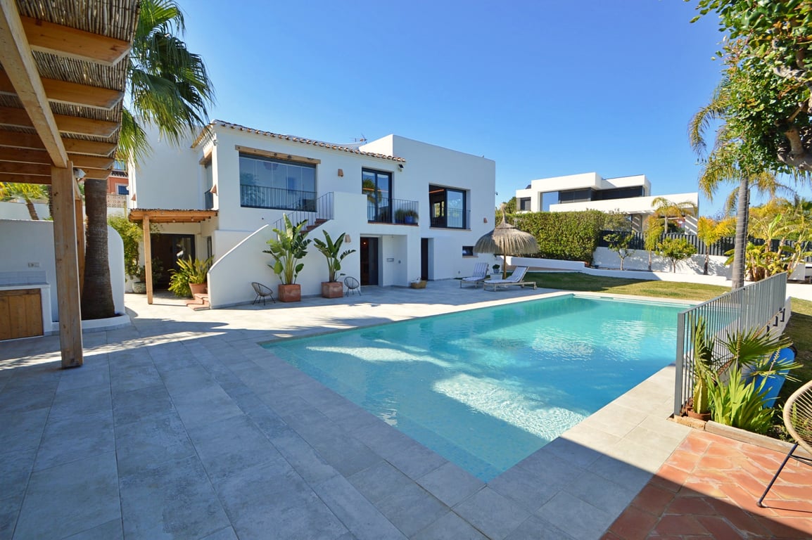 Vue extérieure de la villa avec piscine, terrasse en pierre claire, pergola en bois et façade blanche de style ibicenco avec une naya vitrée à l'étage.