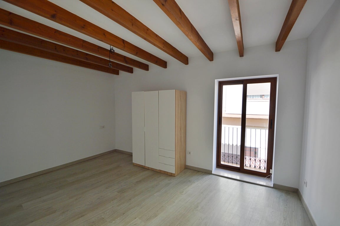 Chambre avec poutres en bois apparentes, sol en stratifié clair et armoire moderne. Une porte vitrée coulissante donne accès à un balcon privé avec garde-corps.
