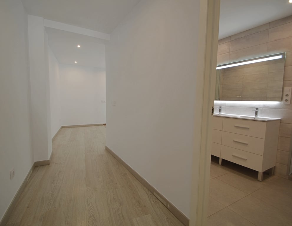 Couloir avec sol en bois clair menant à une salle de bain moderne dotée d'un double lavabo, d'un miroir rétroéclairé et de carrelage mural beige.