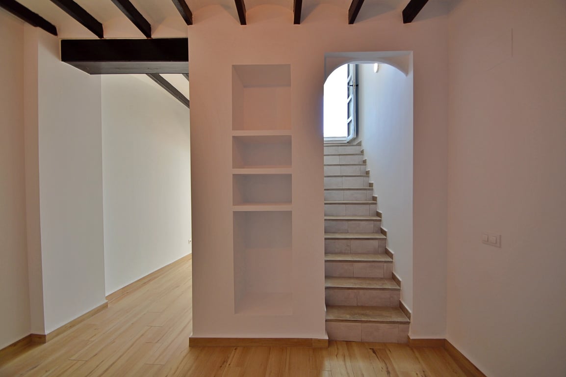 Couloir intérieur avec poutres apparentes en bois sombre, niches murales intégrées et escalier en grès menant à l'étage supérieur baigné de lumière.