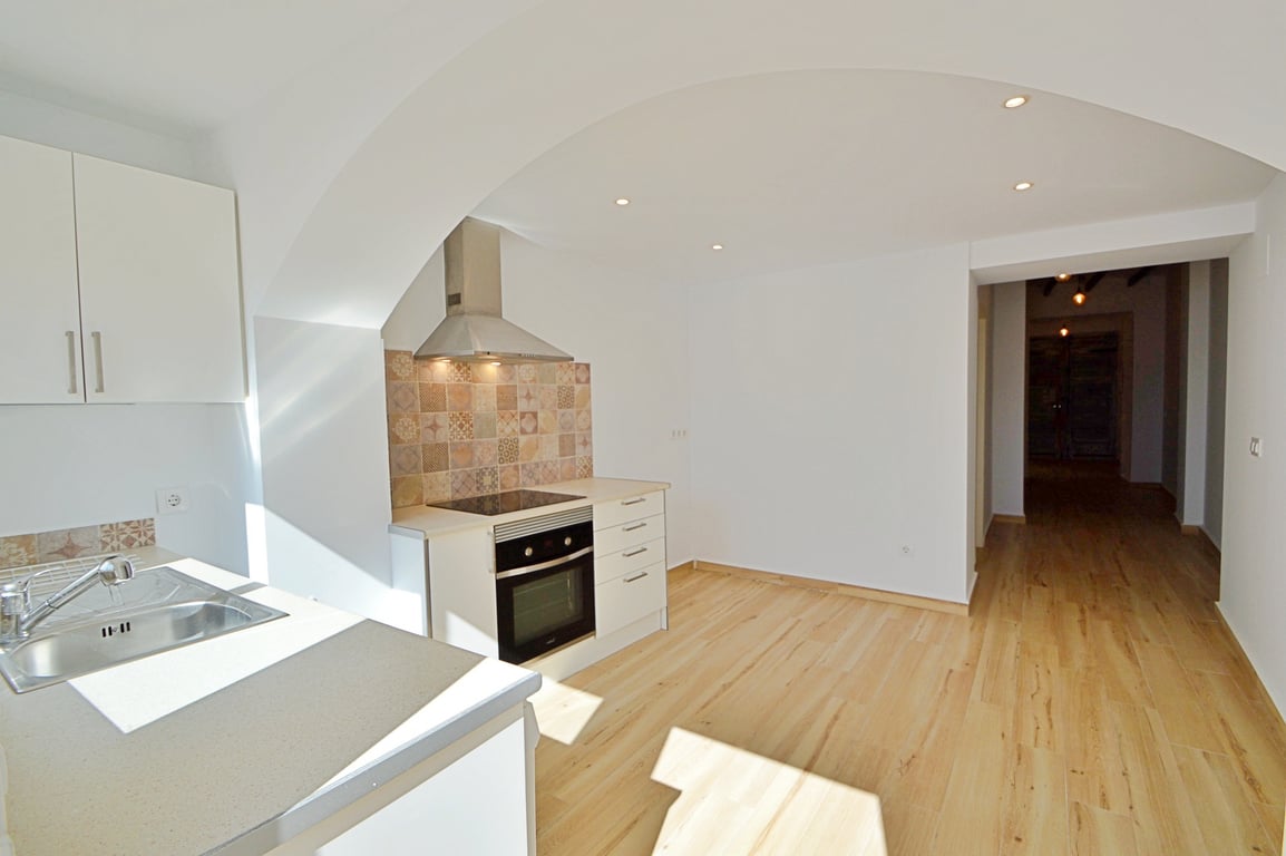Cuisine rénovée avec meubles blancs, crédence en carreaux décoratifs et électroménager intégré. Sol en bois clair, spots encastrés et arche architecturale ouverte.