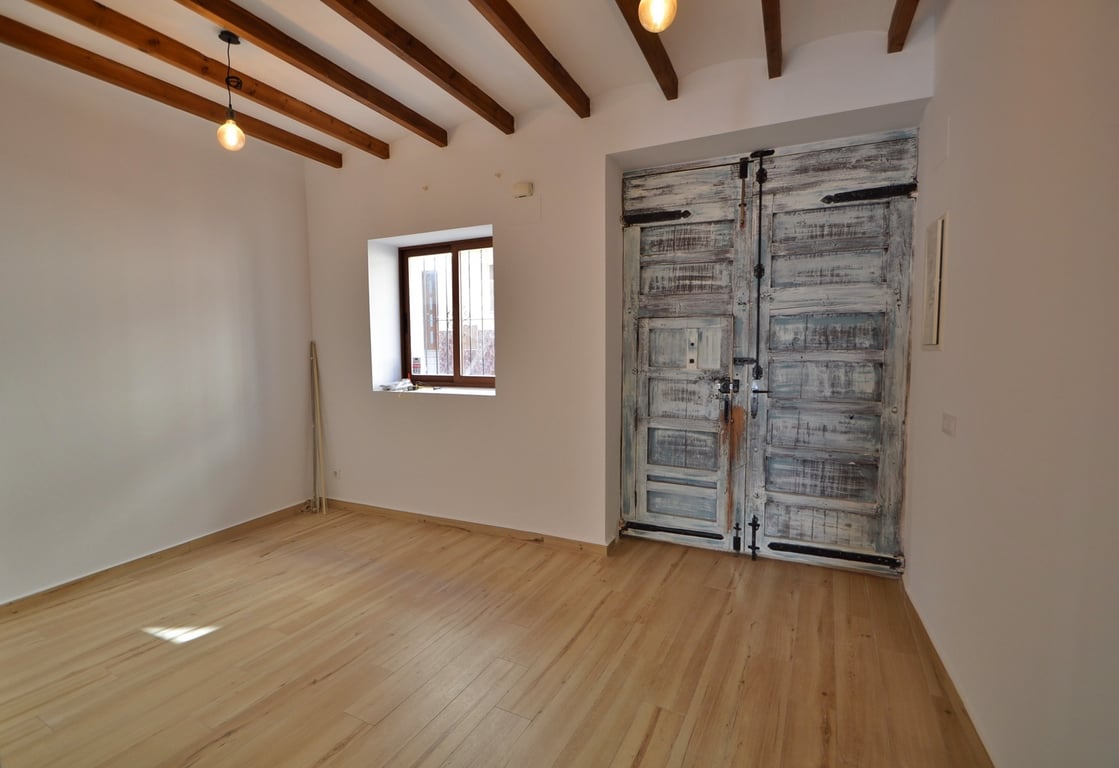 Entrée principale avec double porte en bois rustique restaurée, poutres apparentes et sol en bois clair dans une pièce lumineuse et épurée.