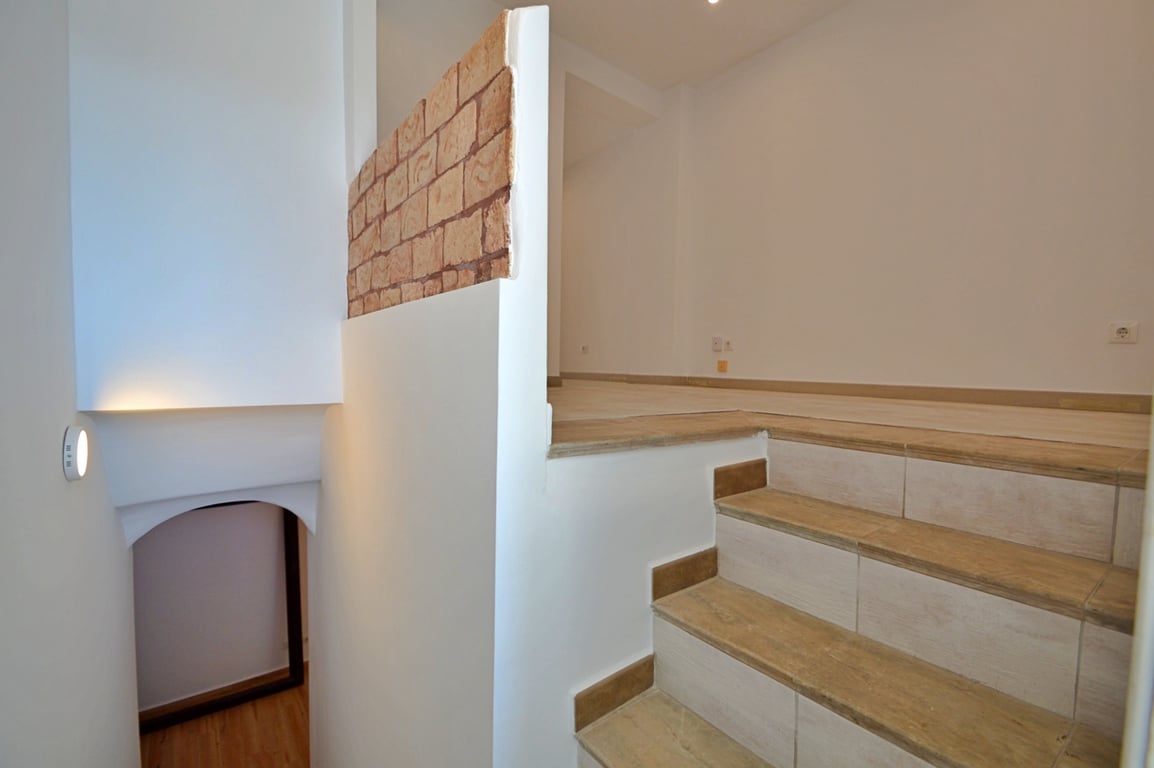 Escalier intérieur avec marches en céramique effet bois, murs blancs et un mur décoratif en briques apparentes séparant les niveaux.