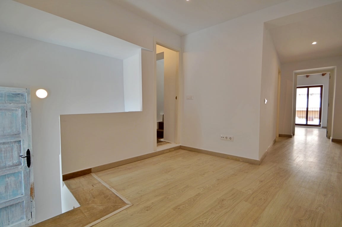 Palier lumineux avec parquet clair, murs blancs et une porte rustique restaurée, menant vers un couloir avec éclairage encastré.