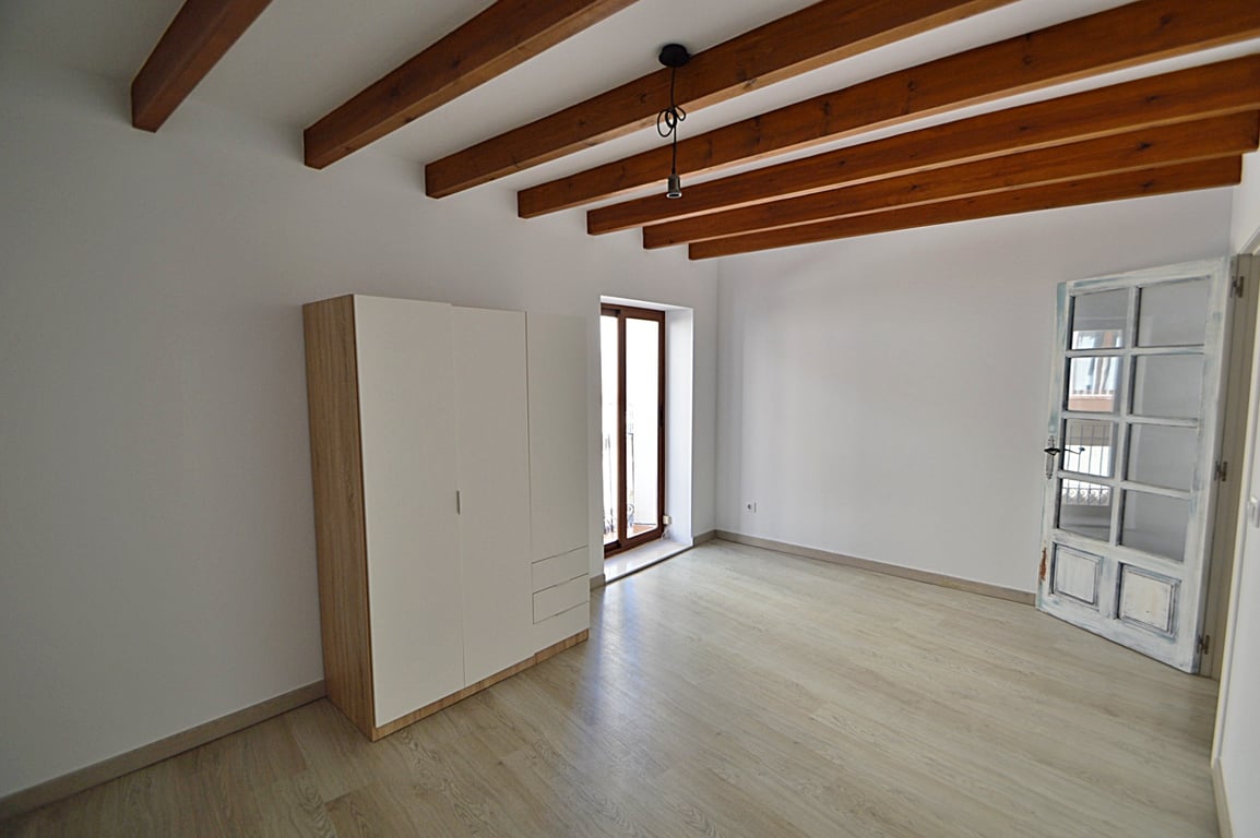 Pièce rénovée avec poutres en bois d'origine, parquet clair et accès au balcon. Comprend une armoire moderne et une porte artisanale en bois avec vitrage.