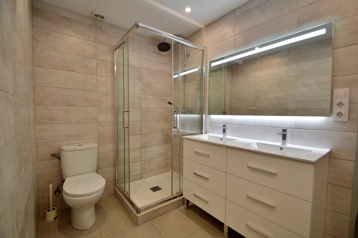 Salle de bain moderne avec double vasque, meuble blanc, grand miroir rétroéclairé et cabine de douche d'angle vitrée sur carrelage beige.