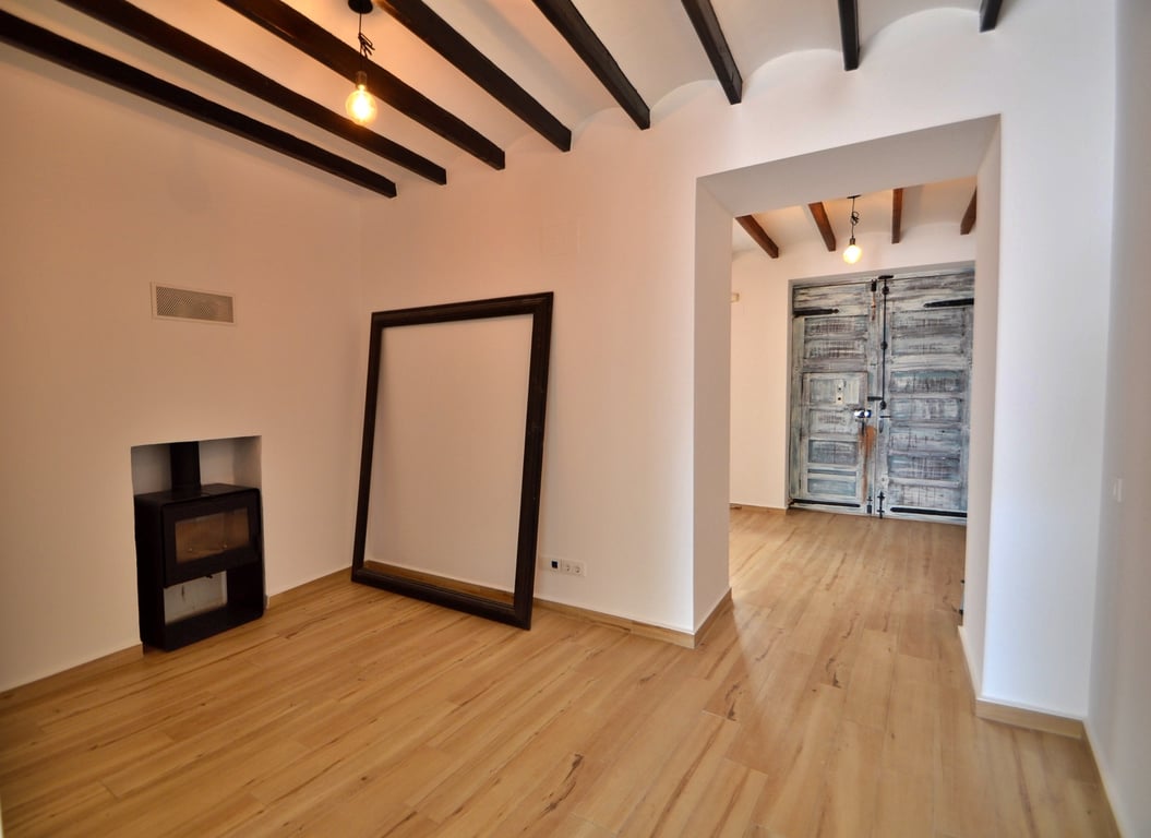 Salon avec poutres apparentes, poêle à bois encastré et sols clairs. Une porte rustique restaurée apporte un cachet authentique à l'espace de vie.