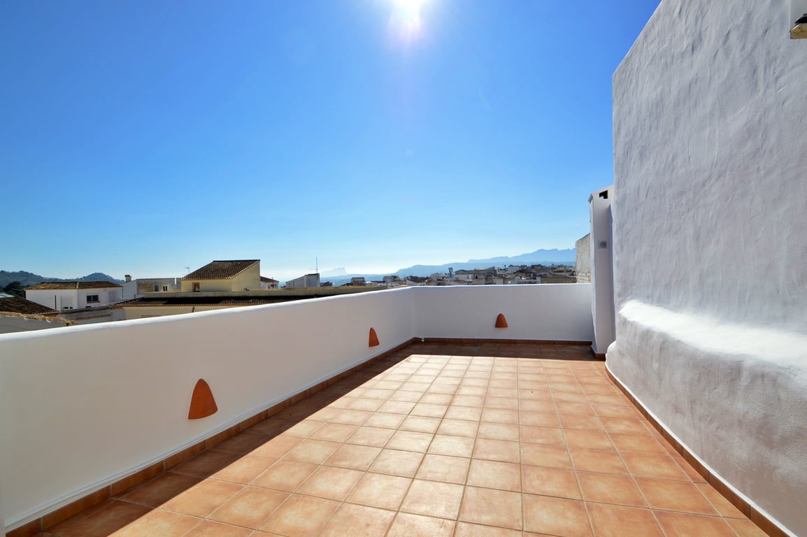 Terrasse sur le toit avec carrelage en terre cuite et murs blancs, offrant une vue panoramique sur les toits du village et les montagnes environnantes.