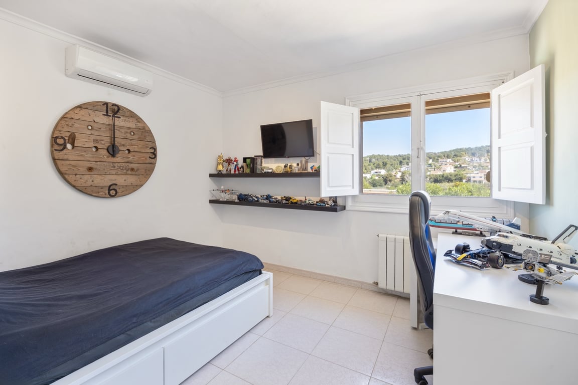 Chambre lumineuse avec climatisation, coin bureau et fenêtre à volets blancs offrant une vue dégagée sur le paysage environnant.