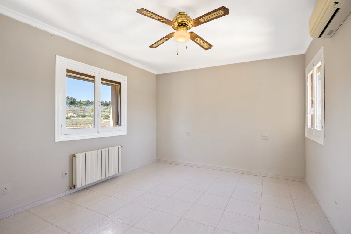 Chambre lumineuse avec sol en grès, ventilateur de plafond, climatisation et radiateur. Les fenêtres offrent une vue dégagée sur le paysage méditerranéen environnant.