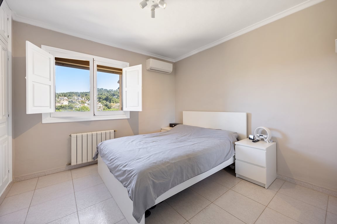 Chambre lumineuse avec volets intérieurs blancs, climatisation et vue dégagée sur les collines. Décoration aux tons neutres avec sol en grès cérame.