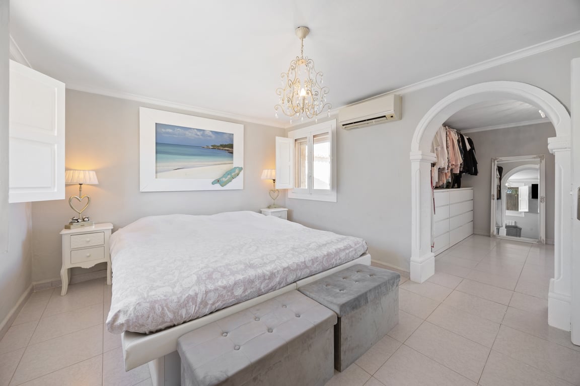 Chambre principale avec dressing attenant via une arche élégante, carrelage clair, climatisation et fenêtres avec volets intérieurs en bois blanc.