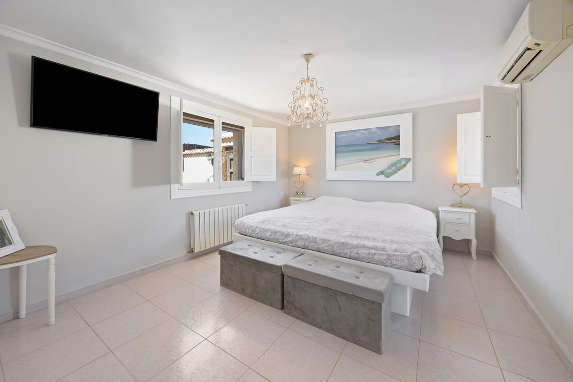 Chambre spacieuse avec volets blancs, télévision murale et climatisation. Elle dispose d'un grand lit avec bancs rembourrés et d'un sol en carrelage clair.