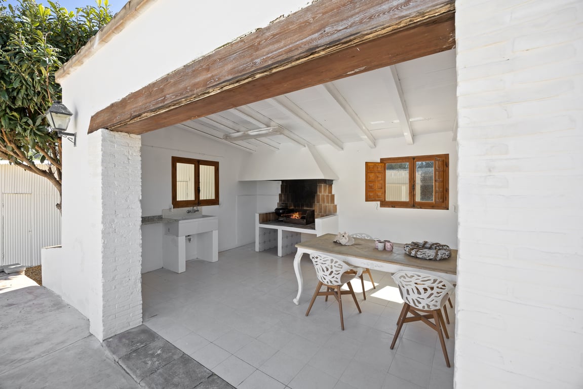 Cuisine d'été couverte avec barbecue intégré, évier en pierre et coin repas, le tout abrité par un plafond à poutres blanches et un linteau en bois massif.