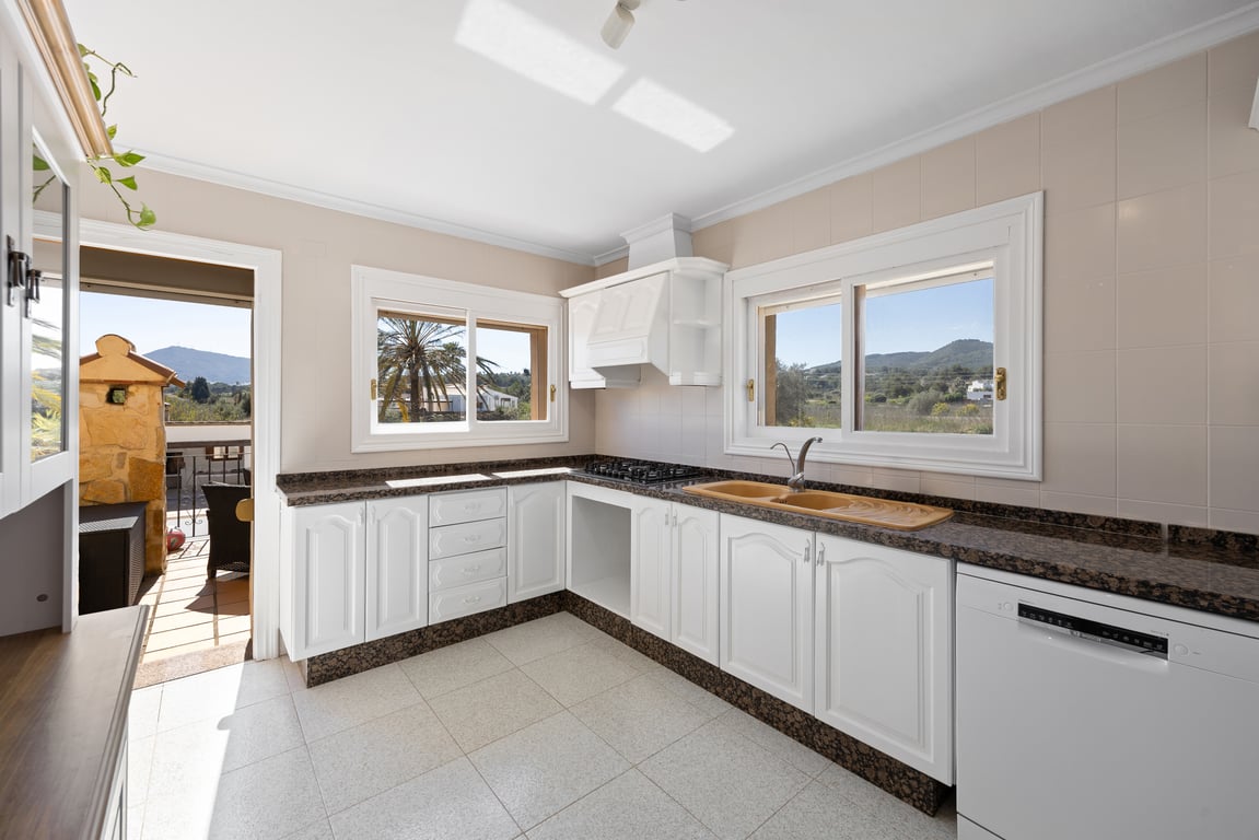 Cuisine lumineuse avec meubles blancs, plans de travail en granit sombre et vue sur la campagne. Accès direct à la terrasse et finitions traditionnelles.