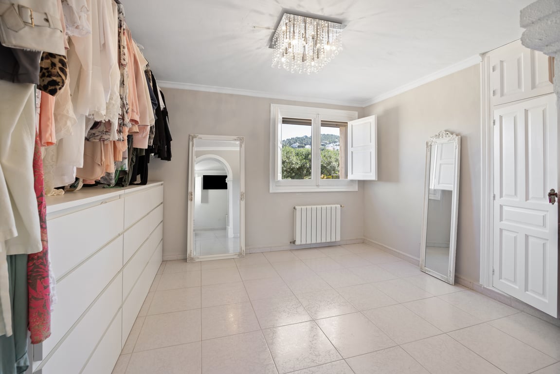 Dressing lumineux avec commode blanche, miroirs sur pied et lustre en cristal. La pièce dispose du chauffage central et d'une fenêtre ouvrant sur la végétation environnante.