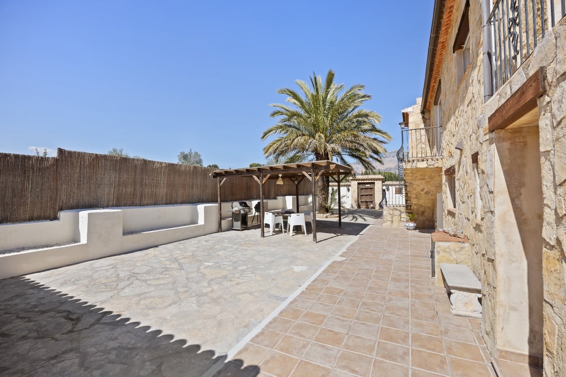 Grande terrasse extérieure avec pavage en pierre, pergola en bois abritant un coin repas et barbecue, bordée par une façade en pierre naturelle.
