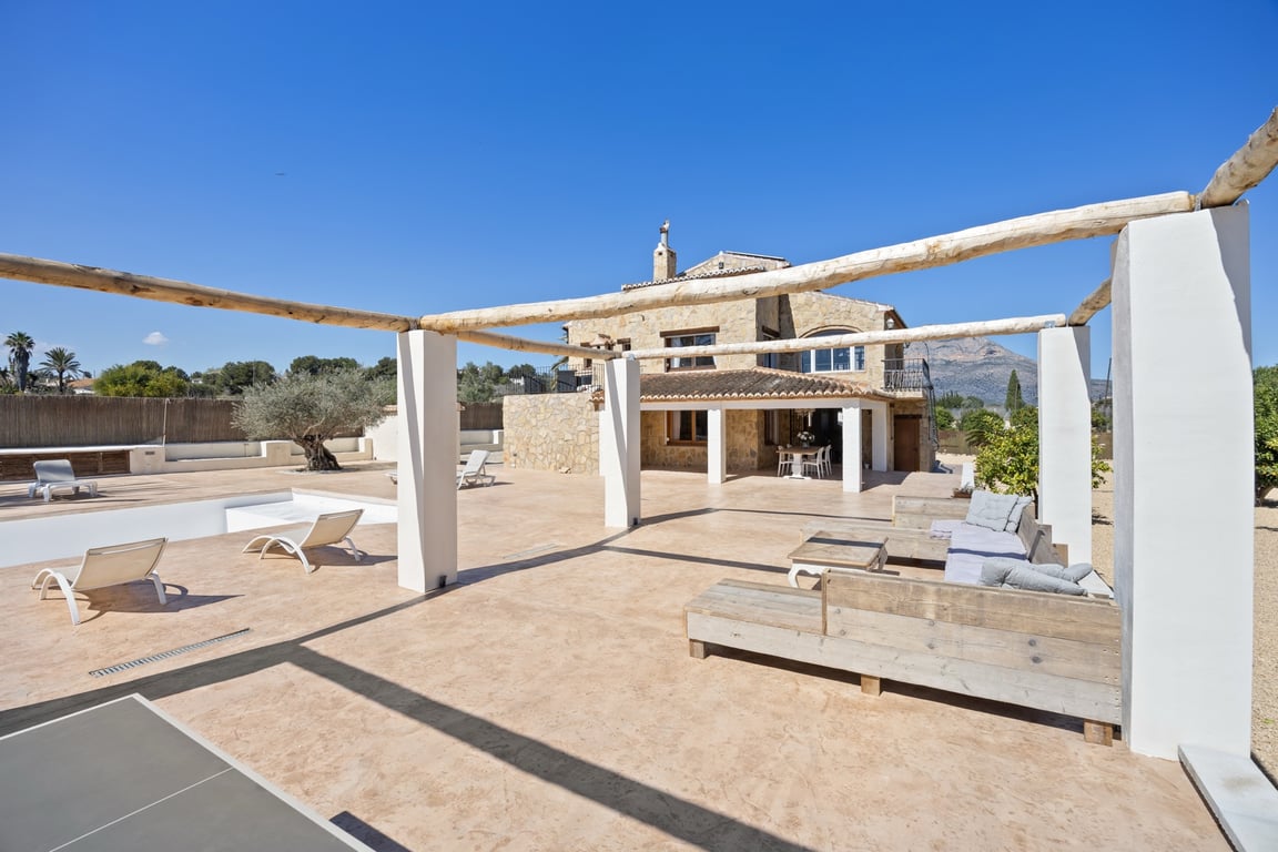 Grande terrasse extérieure avec pergola en poutres de bois, coin salon et vue sur la façade en pierre de la villa et la montagne Montgó.
